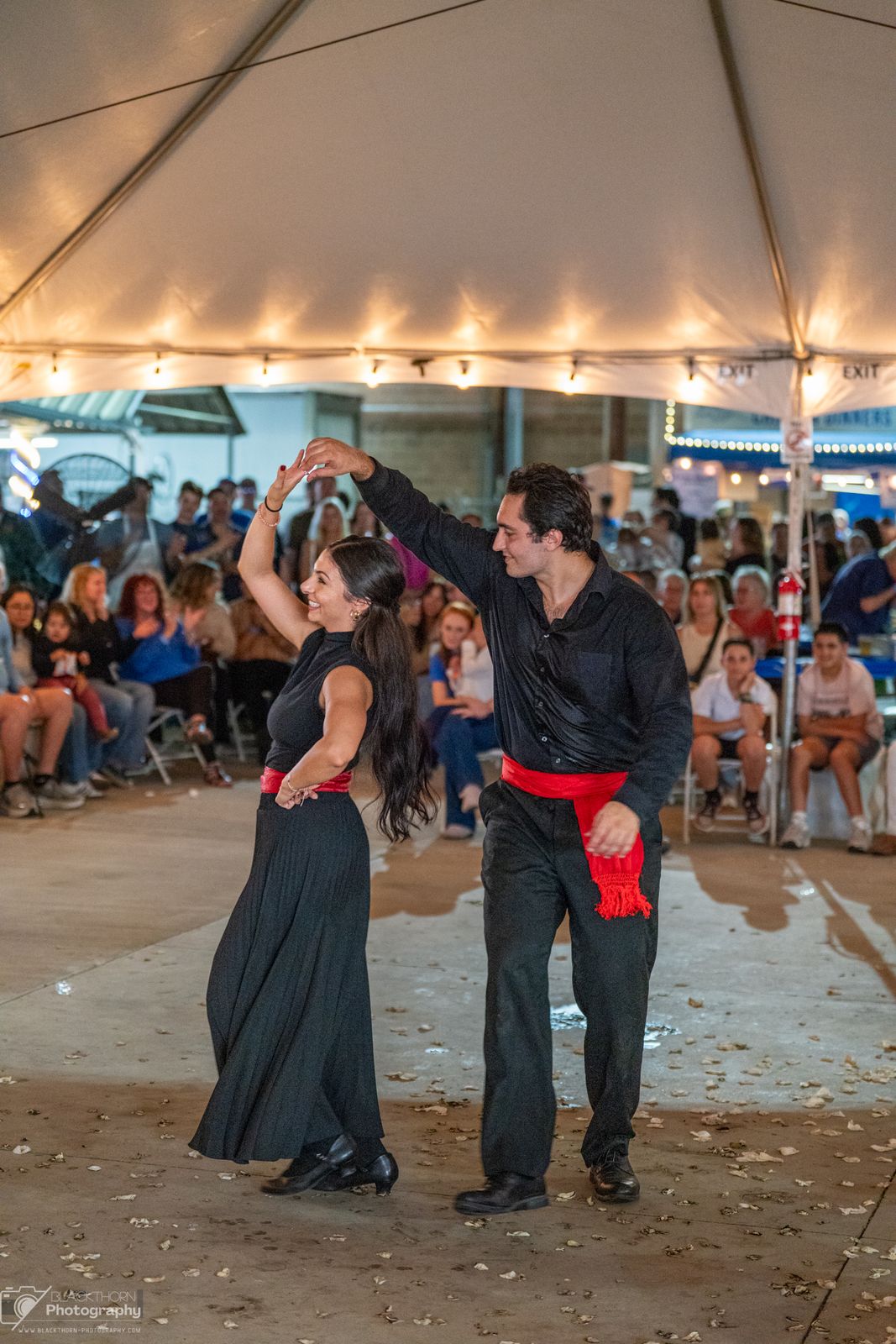 A man and a woman in black with red sashes partner-dancing under the tent