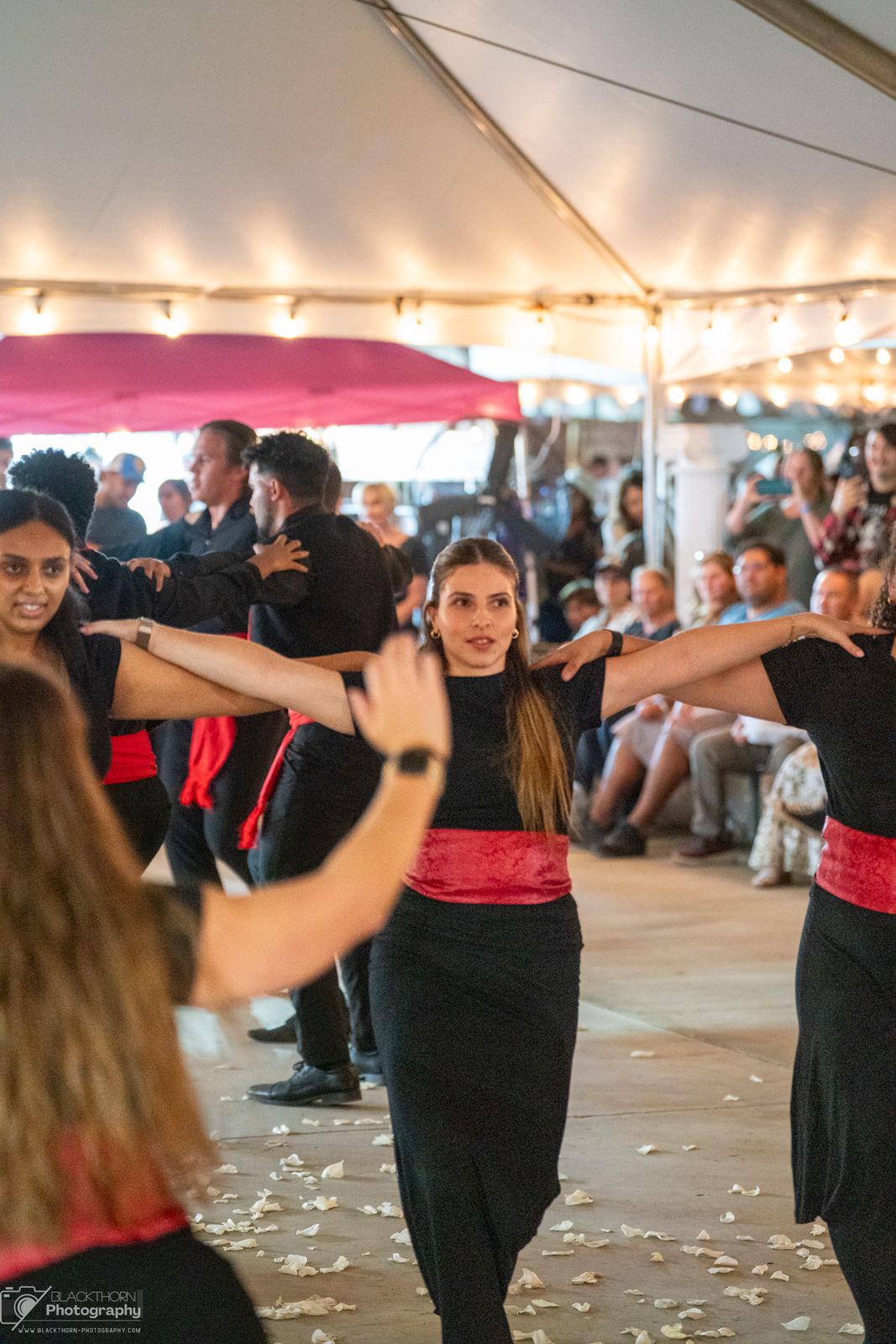 Women in black dresses with red sashes dancing in a linked line under the tent