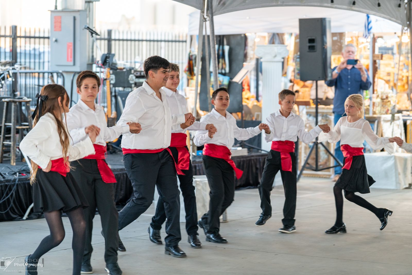 Youth folk dancers in white shirts and red sashes in a line under the tent